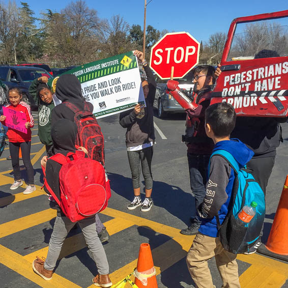 Children walk in crosswalk in front of 'Pedestrians Don't Have Armor' sign