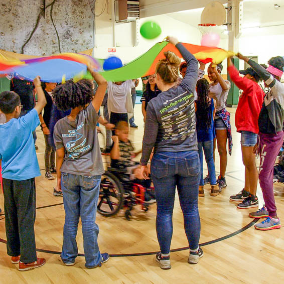 Student using a wheelchair spins under a rainbow parachute held by standing students