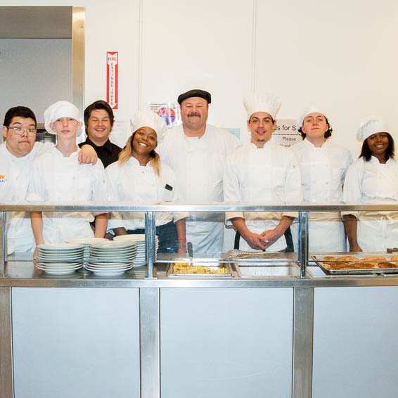 Group of students wearing white chefs hats and aprons, posing with teacher